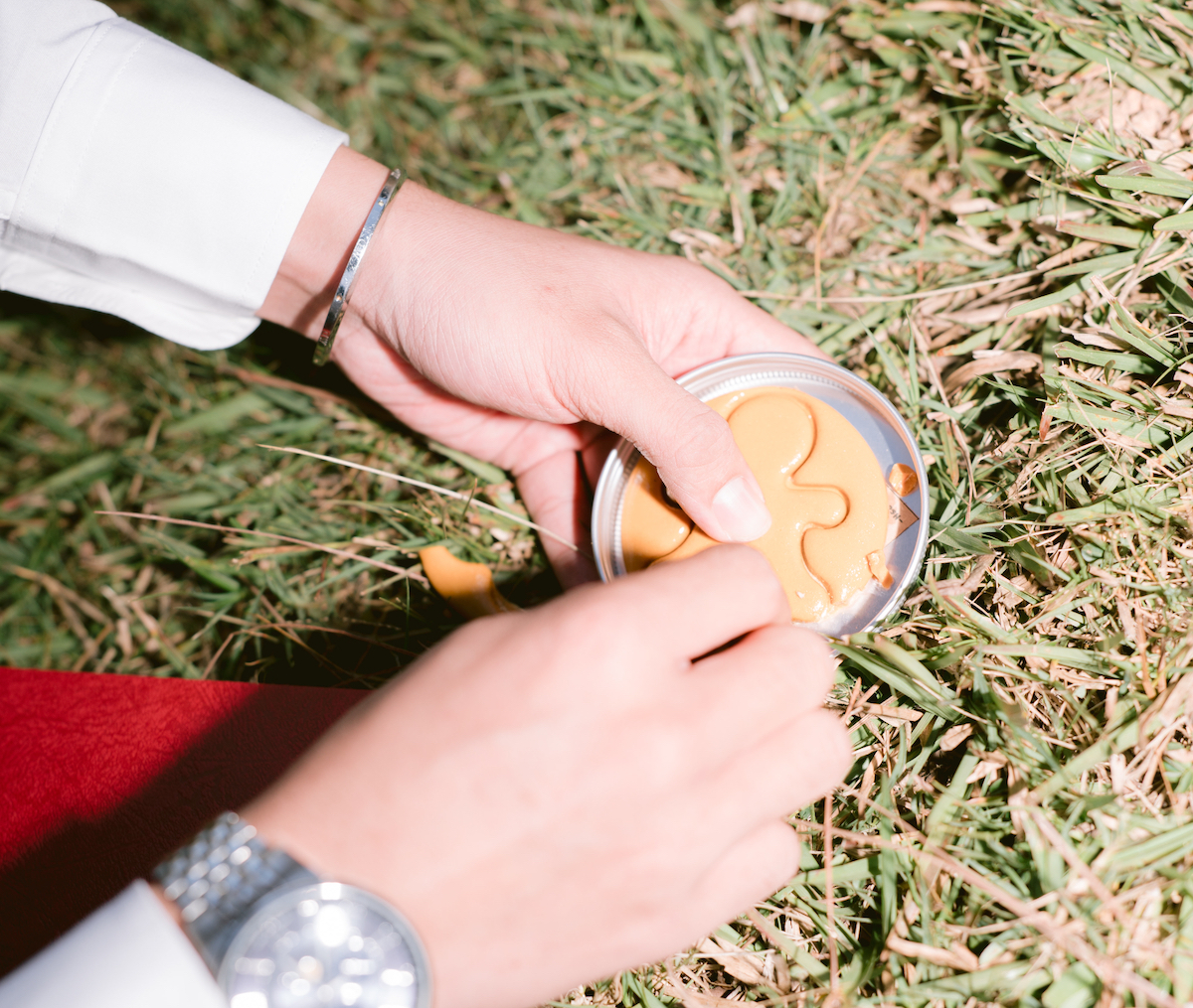 A person uses a tool to cut a shape from a round piece of dalgona candy while sitting on grass.