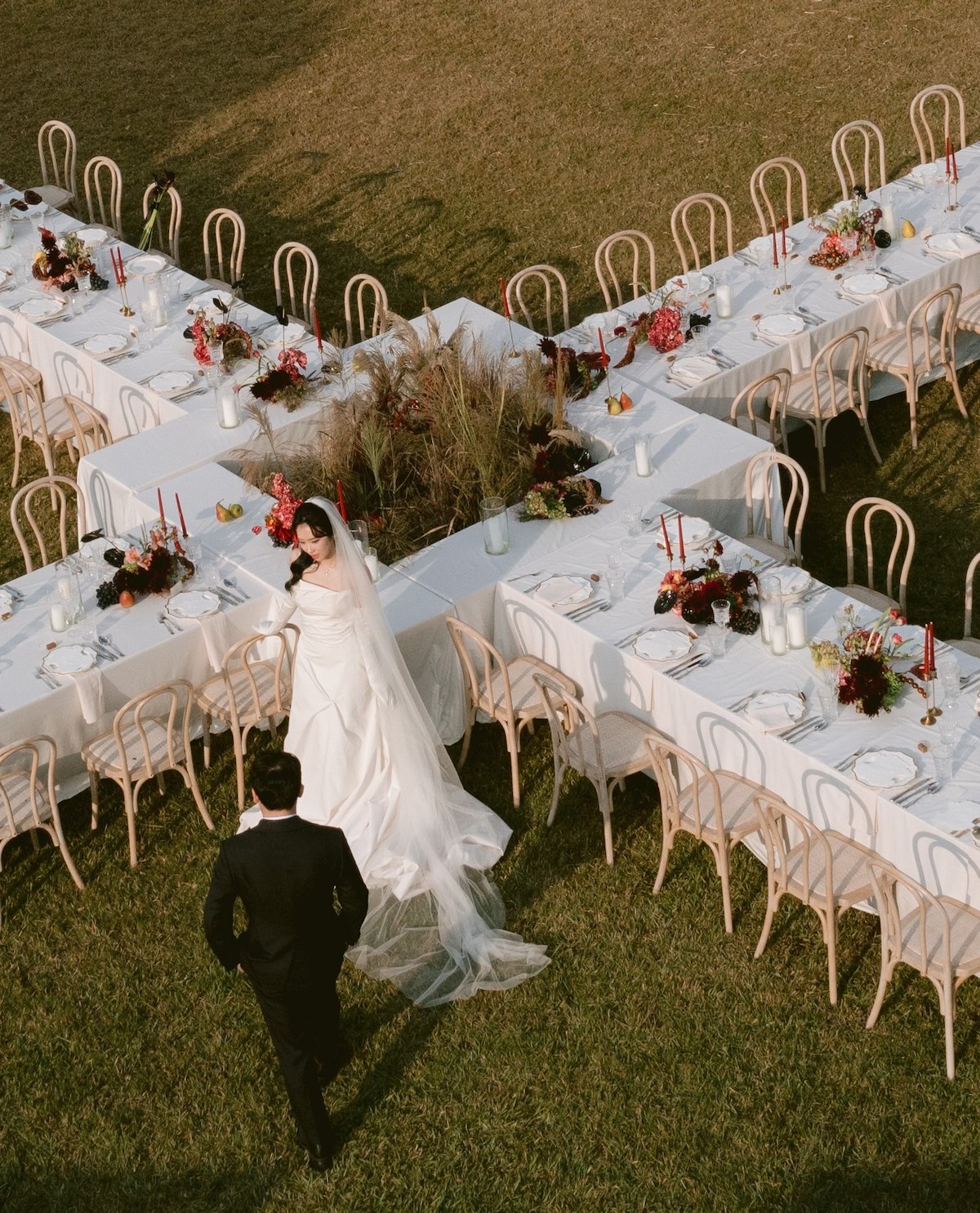 A bride and groom walk beside X-shaped outdoor wedding tables decorated with white tablecloths, flowers, and candles on a grassy lawn.