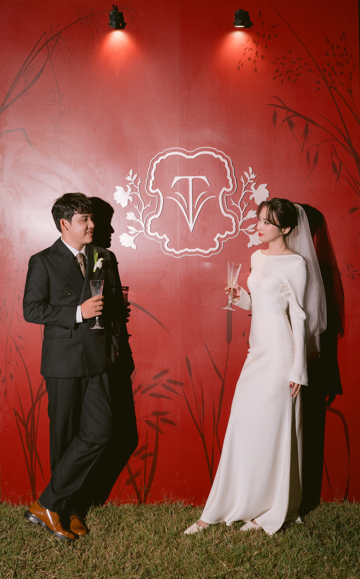 A bride and groom stand in front of a red wall with white floral designs, holding champagne glasses and facing each other.