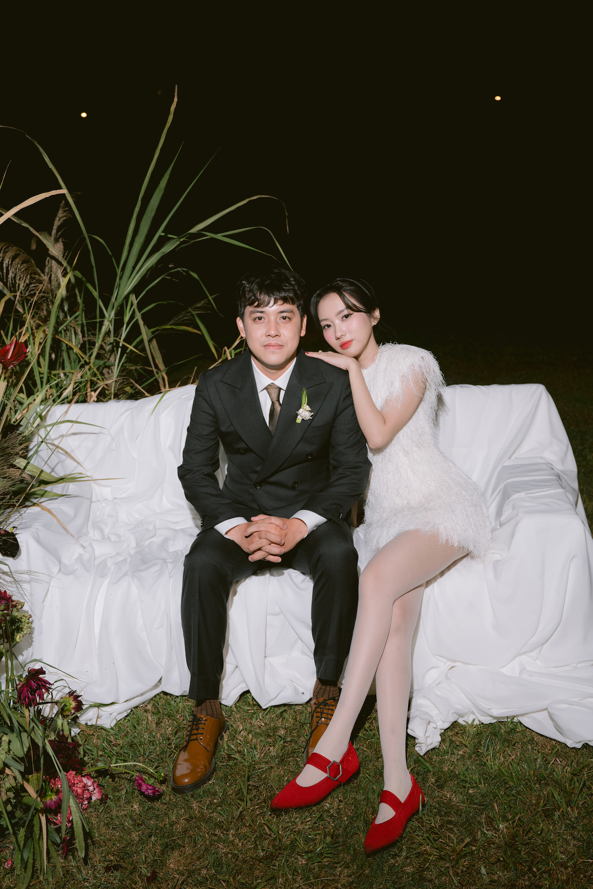 A well-dressed couple sits on a white-covered outdoor sofa at night, with the woman resting her head on the man's shoulder.