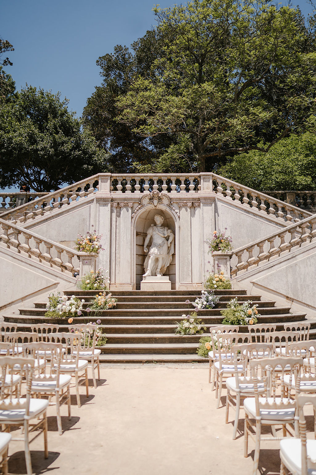 Outdoor stone staircase with balustrades, floral arrangements, a central classical statue in an alcove, and rows of chairs set up for an event.
