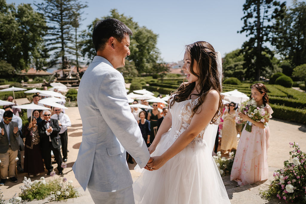 A bride and groom hold hands and smile at each other during an outdoor wedding ceremony, with guests and bridesmaids in the background.