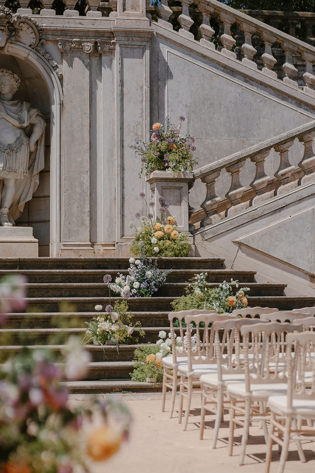 Rows of white chairs face stone steps decorated with flower arrangements, with a large statue and ornate balustrade in the background.