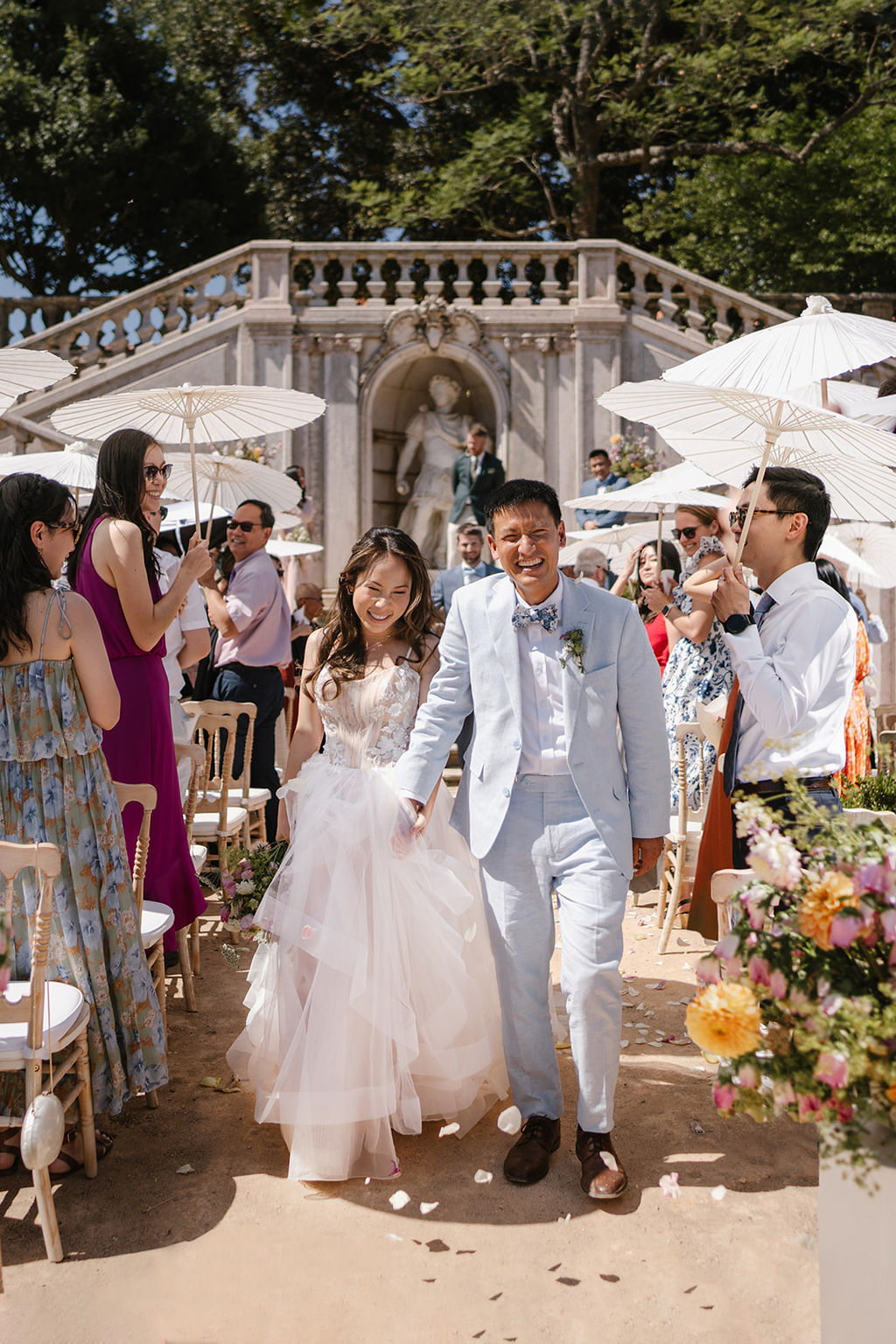 A bride and groom walk down an outdoor aisle smiling, surrounded by guests holding white umbrellas at a daytime wedding ceremony.
