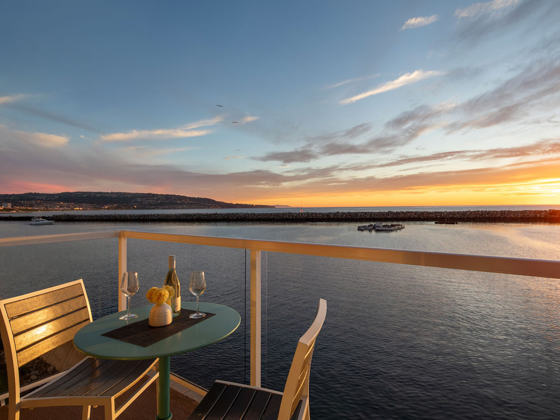 Table with two chairs, a bottle of wine, glasses, and a flower vase on a balcony overlooking calm water and a sunset sky.