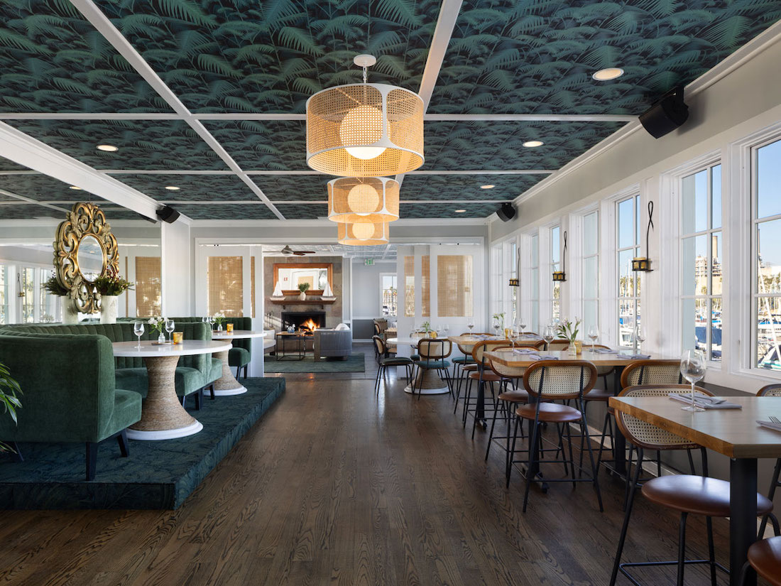 Modern restaurant interior with wooden floors, patterned ceiling, round pendant lights, green velvet seating, and large windows letting in natural light.