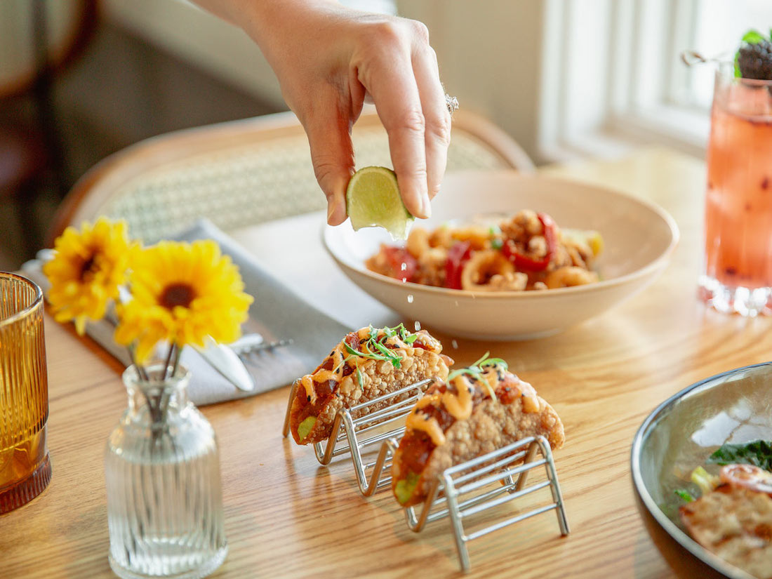 A hand squeezes a lime wedge over tacos on a table set with a bowl of food, a drink, and a small vase of yellow flowers.