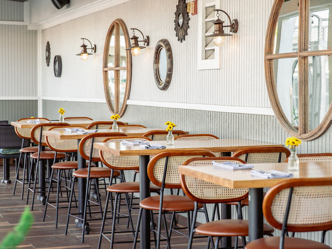A row of tables and chairs in a bright, modern restaurant with round mirrors and wall lamps on a white paneled wall. Small vases with yellow flowers are on each table.