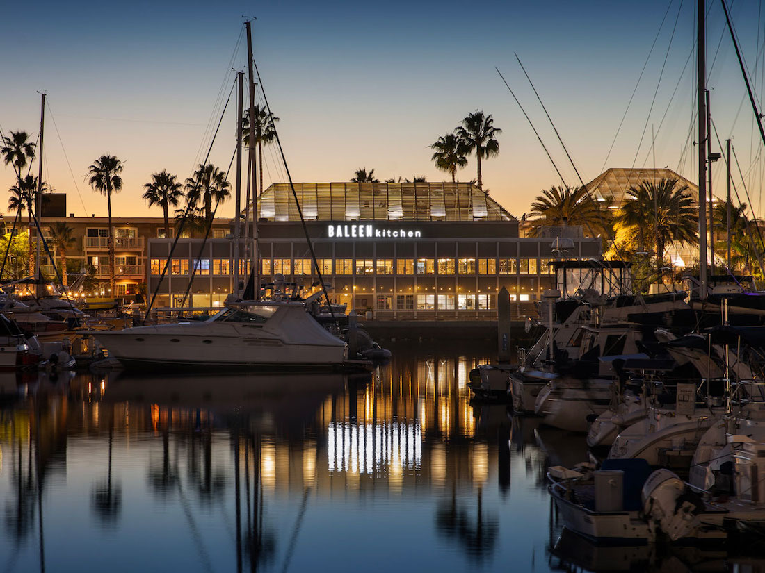 Yachts docked in a harbor at dusk with a building labeled "Baleen Kitchen" illuminated in the background. Palm trees are silhouetted against the evening sky.