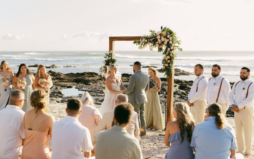 A couple stands under a floral arch exchanging vows during a beach wedding ceremony, with guests seated and an officiant present.