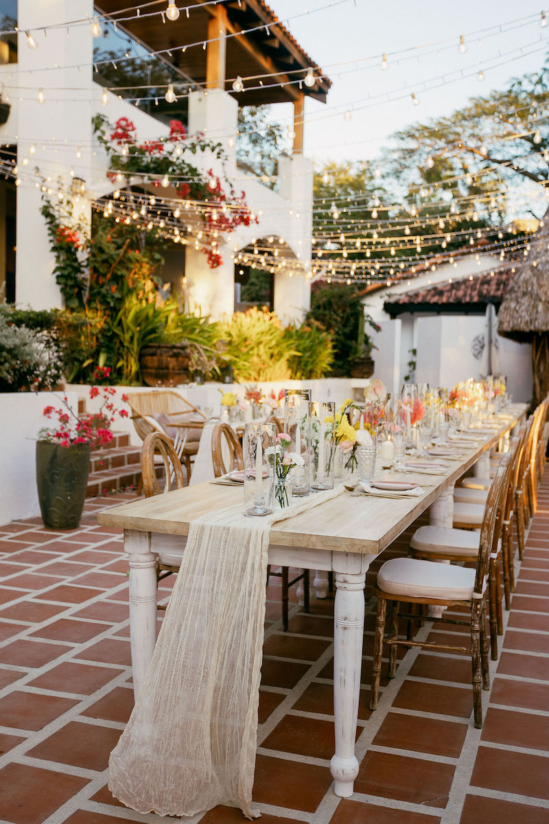 A long outdoor dining table with floral centerpieces is set for a meal on a tiled patio, surrounded by string lights and greenery.