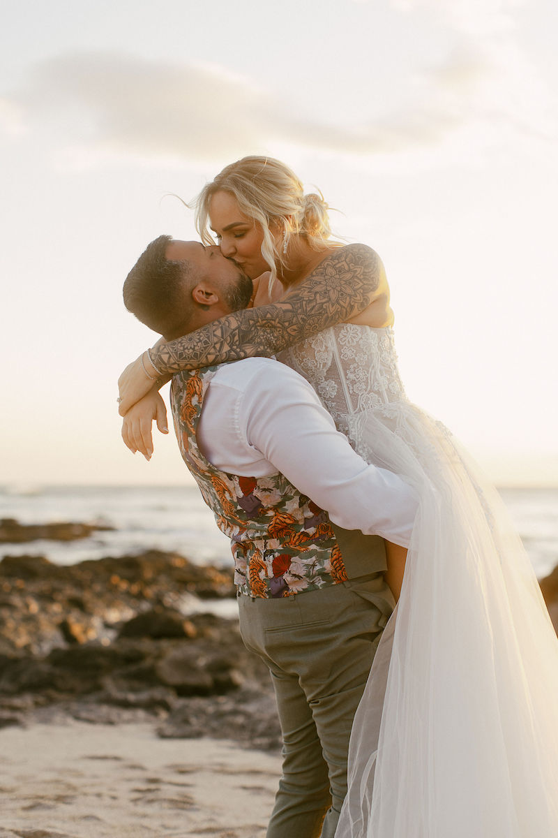 A bride in a white dress is lifted and embraced by a groom in a floral vest on a sandy beach at sunset.