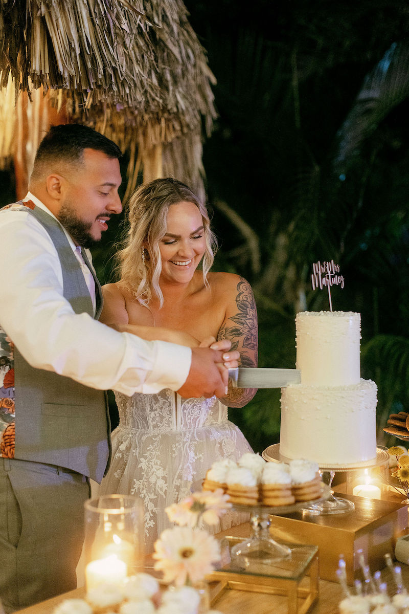A bride and groom cut a white tiered wedding cake together at a decorated outdoor reception table, surrounded by candles and cupcakes.