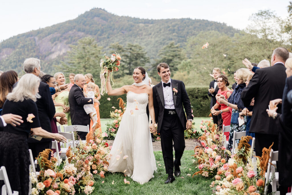 A bride and groom walk down an outdoor aisle lined with flowers, smiling and holding hands, as guests celebrate around them with flower petals. Mountains and trees are visible in the background.