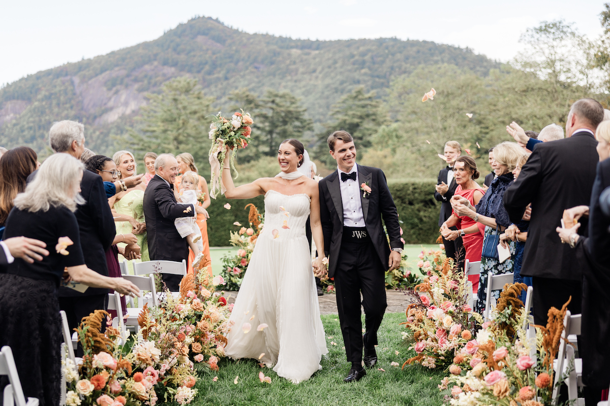 A bride and groom walk down an outdoor aisle lined with flowers, smiling and holding hands, as guests celebrate around them with flower petals. Mountains and trees are visible in the background.