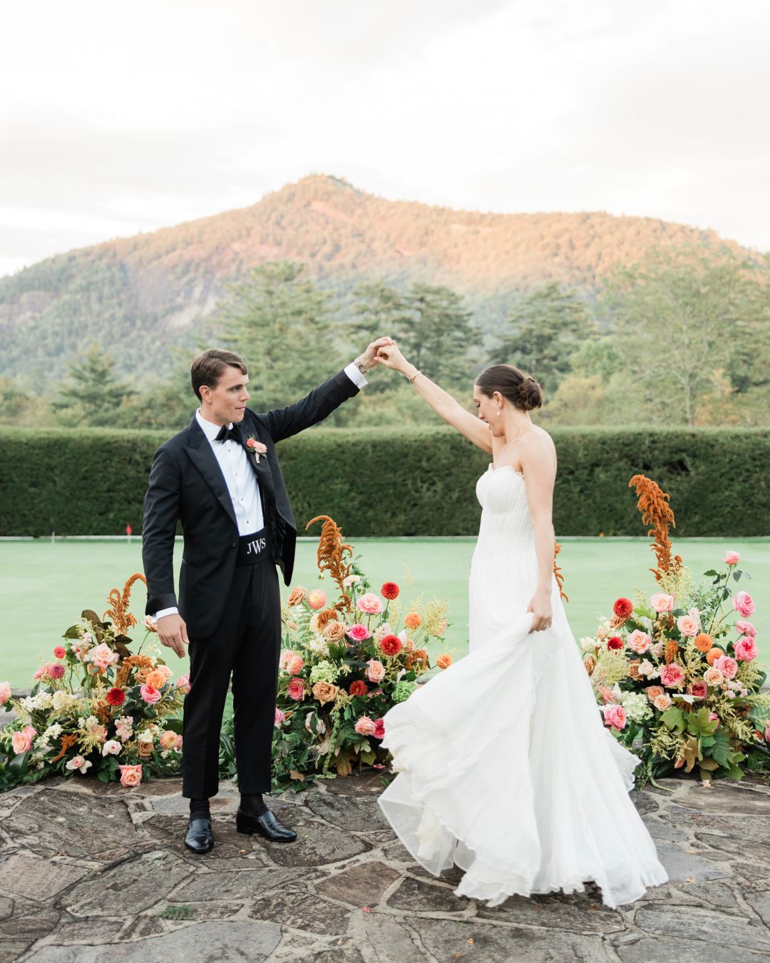 A bride and groom dance outdoors in front of a flower arrangement, with a mountain and greenery in the background.