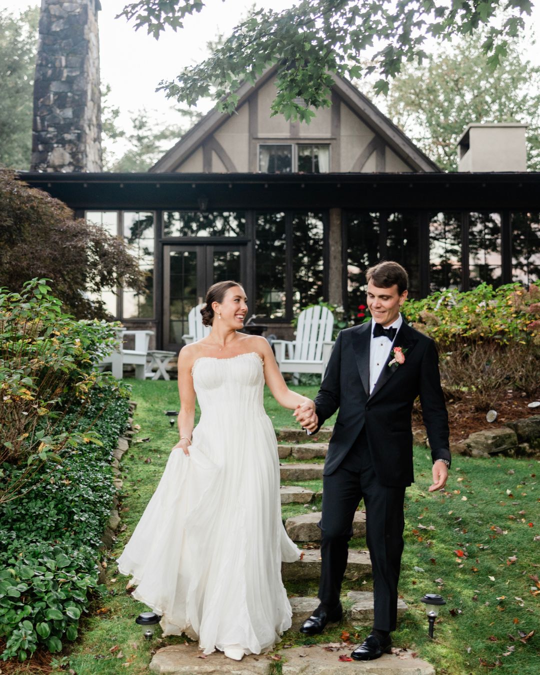 A bride in a white gown and a groom in a black tuxedo walk hand in hand down stone steps in front of a house with greenery around them.