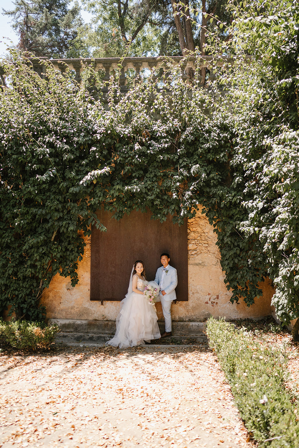 A couple stands together in front of a large wooden door framed by dense green ivy on a sunny day.