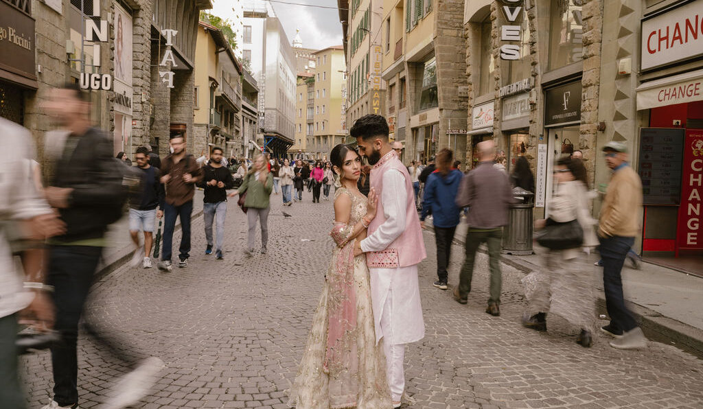 A couple in traditional South Asian attire stands together on a busy cobblestone street, surrounded by blurred pedestrians and historic buildings.