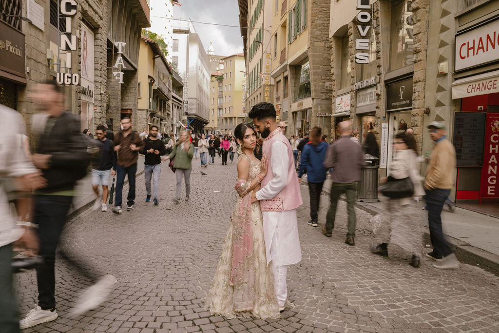 A couple in traditional South Asian attire stands together on a busy cobblestone street, surrounded by blurred pedestrians and historic buildings.