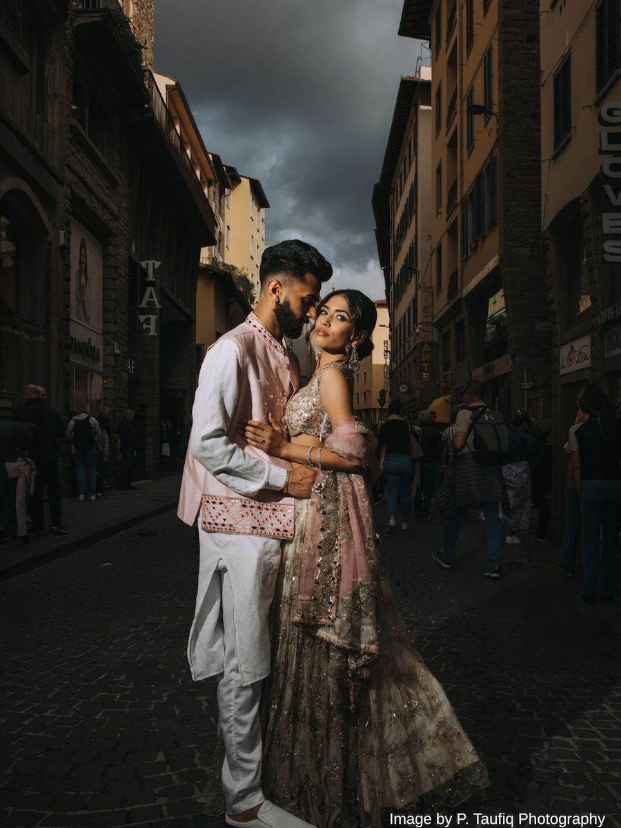 A couple in formal South Asian attire pose closely together on a cobblestone street in Italy, with dramatic lighting and cloudy skies above, capturing the elegance of a destination wedding.