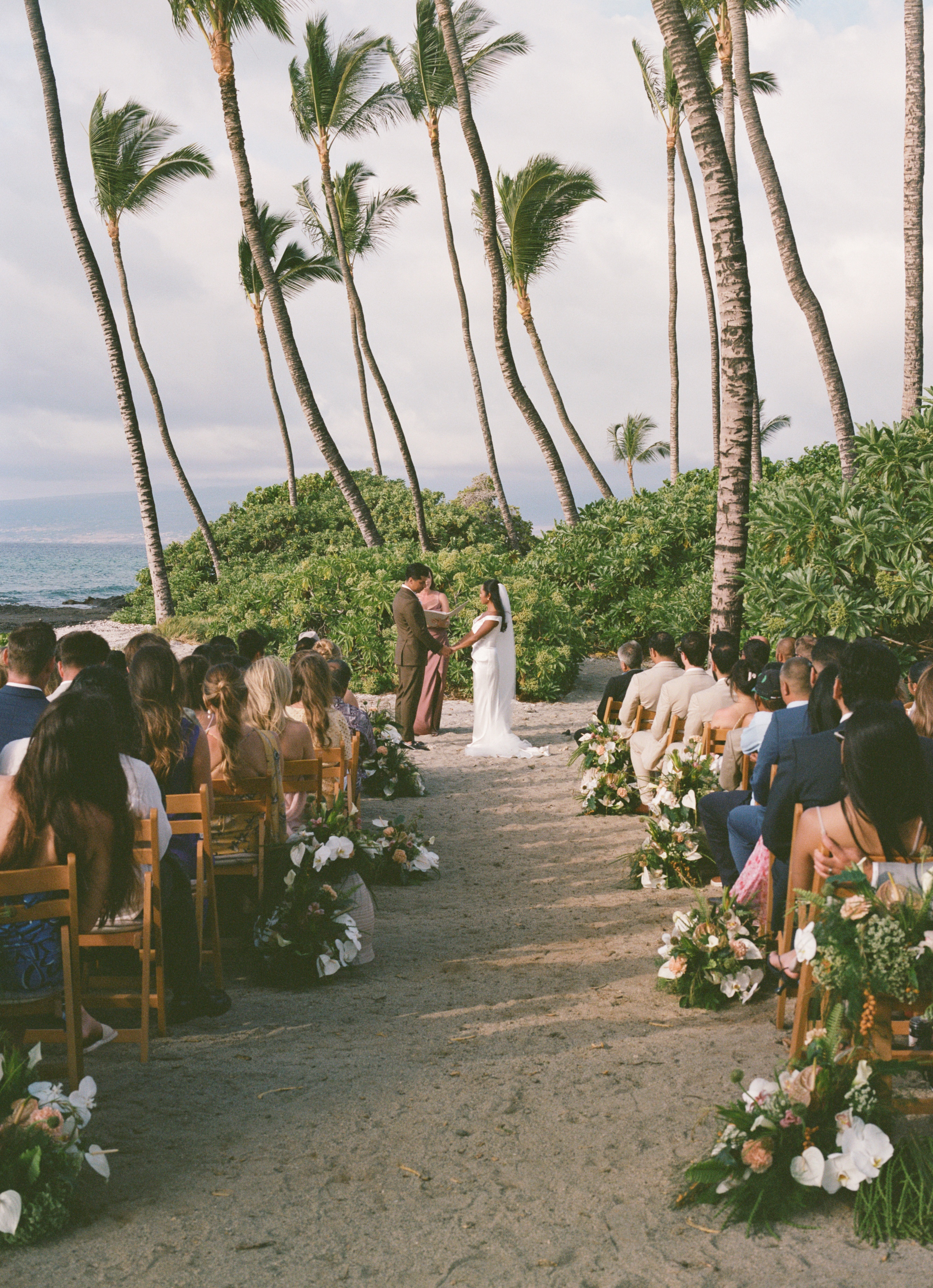 A bride and groom stand with an officiant during an outdoor beach wedding ceremony, surrounded by guests seated along an aisle lined with floral arrangements and palm trees.