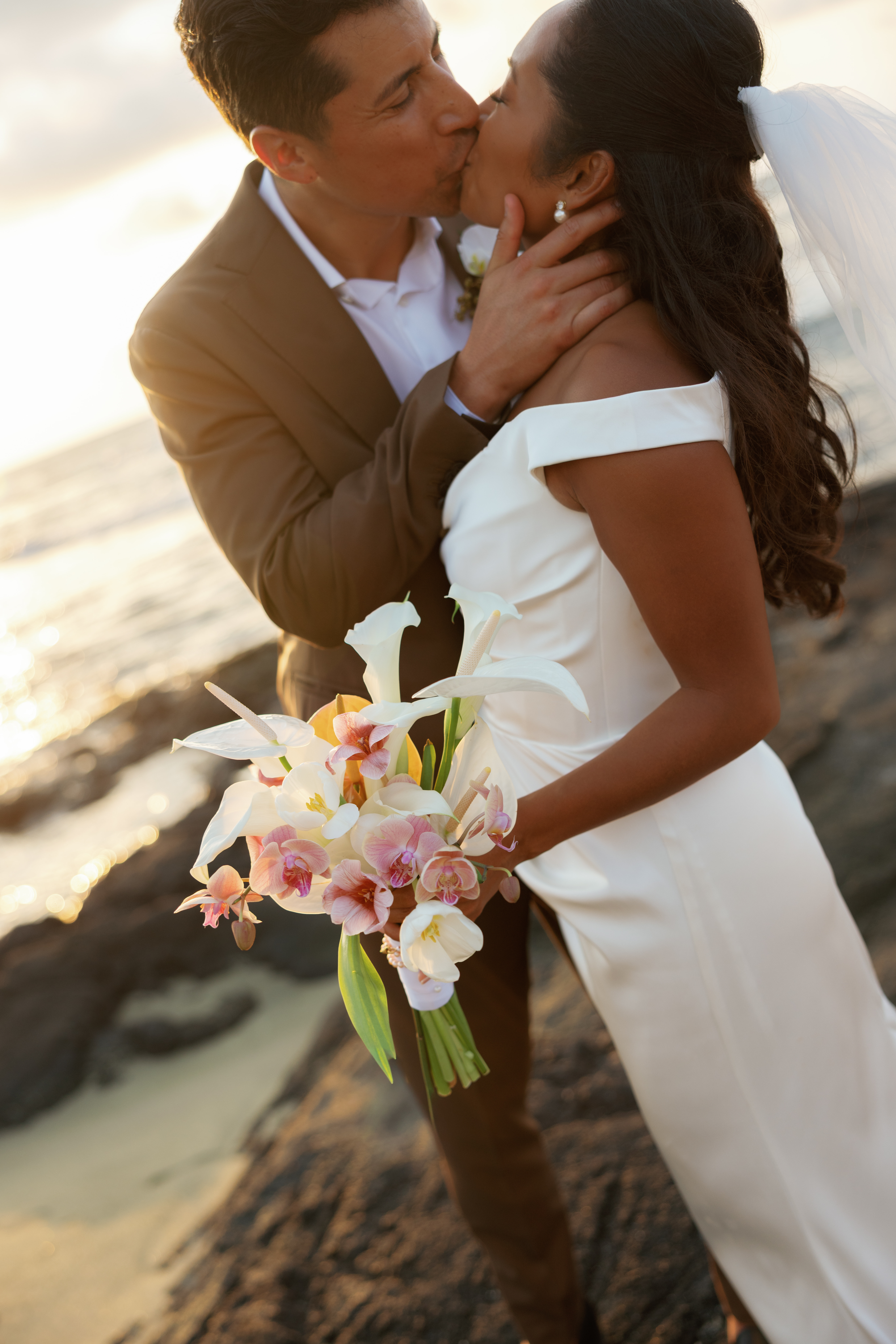 A bride and groom kiss on a beach at sunset; the bride holds a bouquet of white and pink flowers.