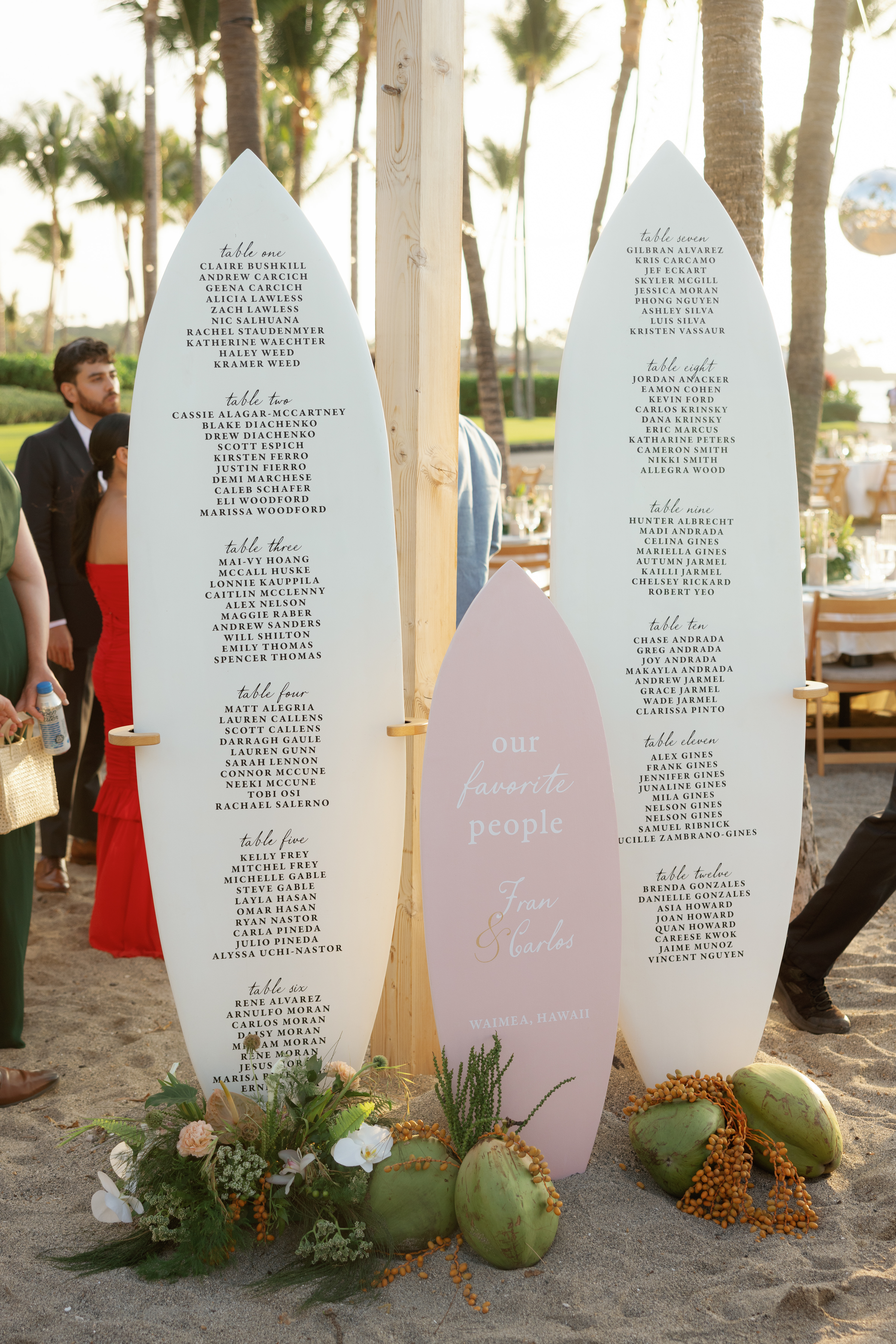 Three surfboards, two white and one pink, display seating arrangements at an outdoor beach event, with tropical flowers and coconuts at the base.