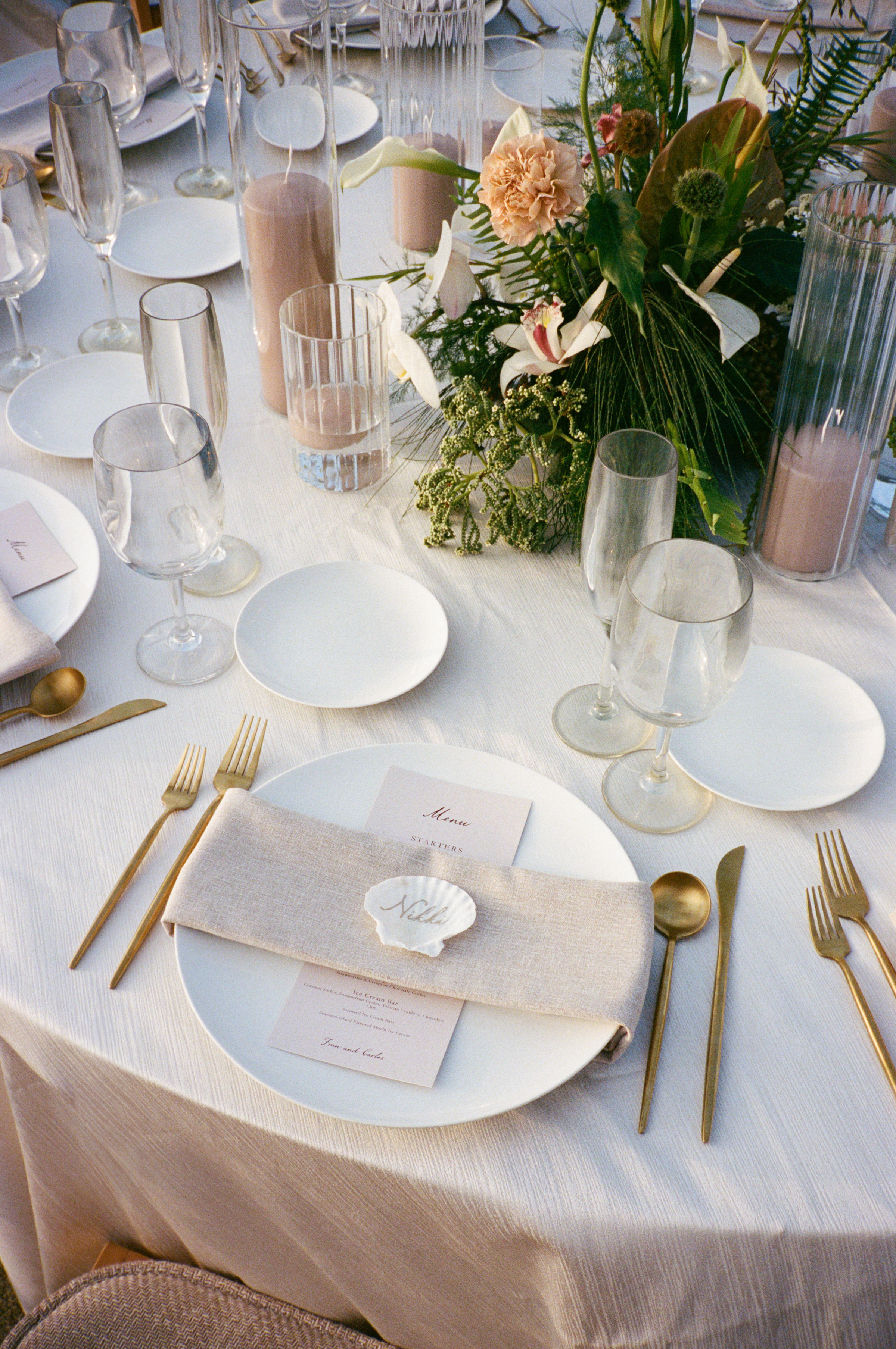 A formal table setting with white plates, gold cutlery, glassware, beige napkins, and a floral centerpiece on a white tablecloth.
