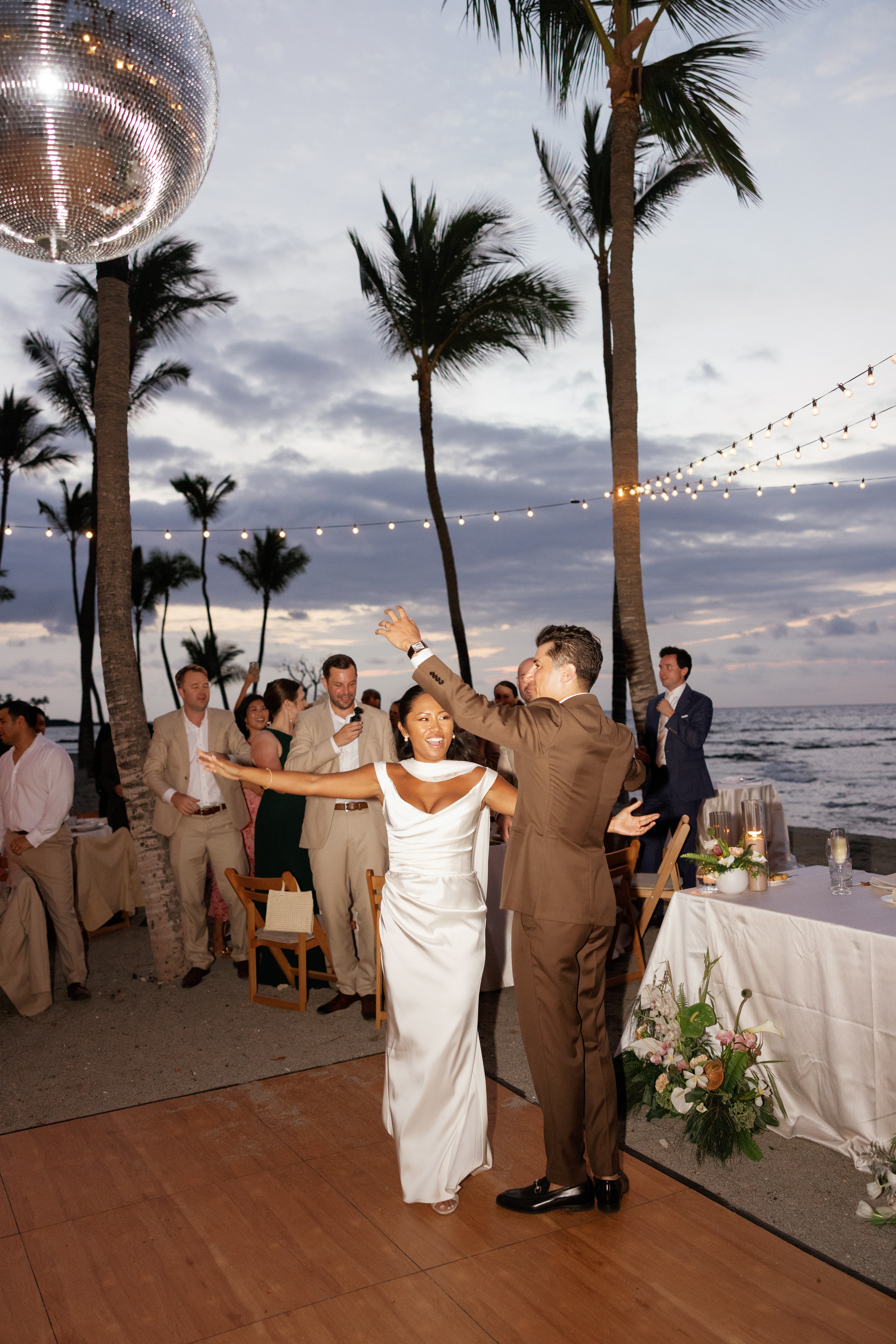A bride and groom dance together on an outdoor floor by the beach at sunset, surrounded by guests, palm trees, string lights, and a large disco ball overhead.