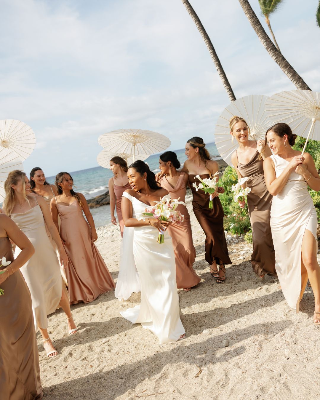 A bride in a white dress walks on a sandy beach with bridesmaids in neutral dresses, holding white umbrellas and bouquets, with the ocean and palm trees in the background.
