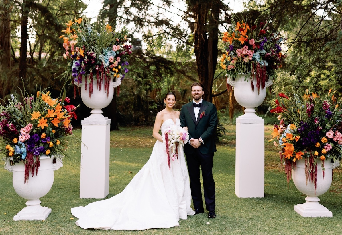 A bride and groom stand outdoors between two large floral arrangements on pillars, posing for a wedding photo.
