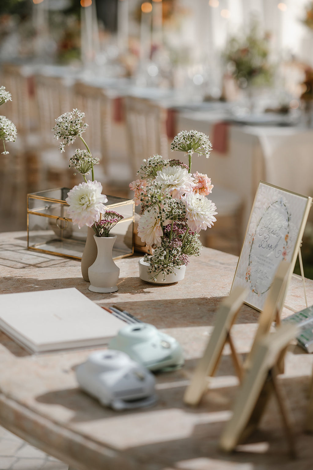 A table decorated with flowers, a guestbook, instant cameras, and a framed sign at an event, with chairs and a decorated table in the background.
