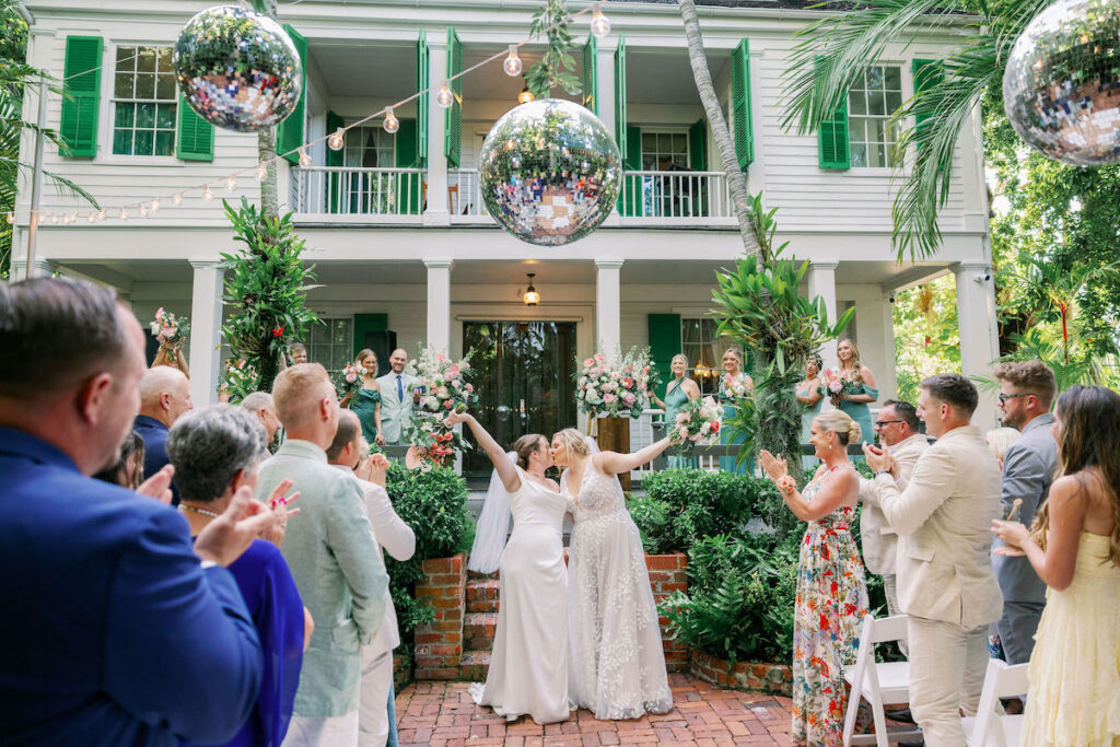 Two brides kiss and raise their arms in celebration at an outdoor wedding ceremony, surrounded by applauding guests, with a large disco ball and greenery in the background.
