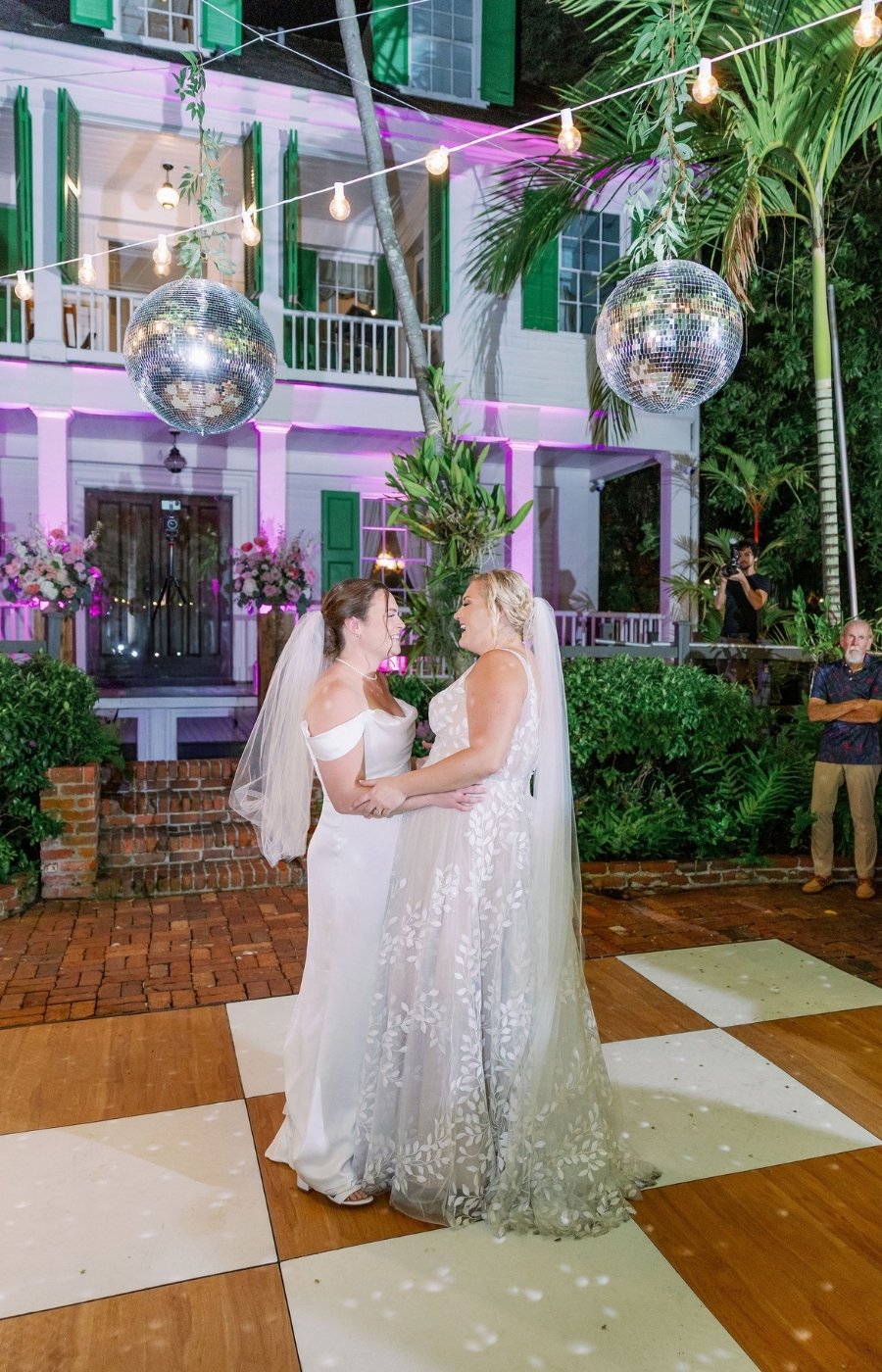 Two brides in wedding dresses hold hands and dance together on a checkered outdoor dance floor, with a house, disco balls, and string lights in the background.