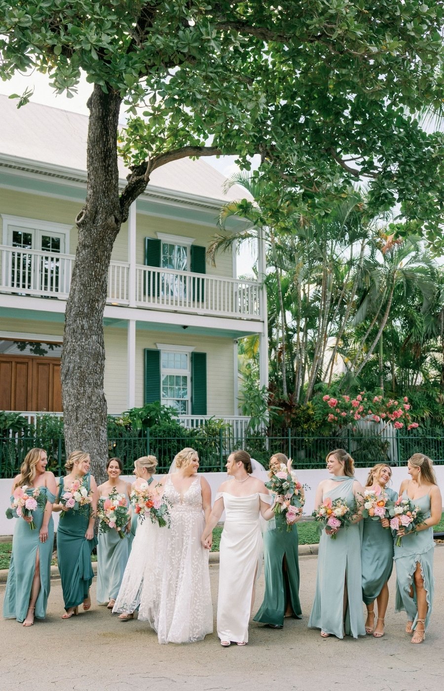 A group of women in light blue and white dresses, holding bouquets, stand together on a street in front of a house with white walls and a large tree.