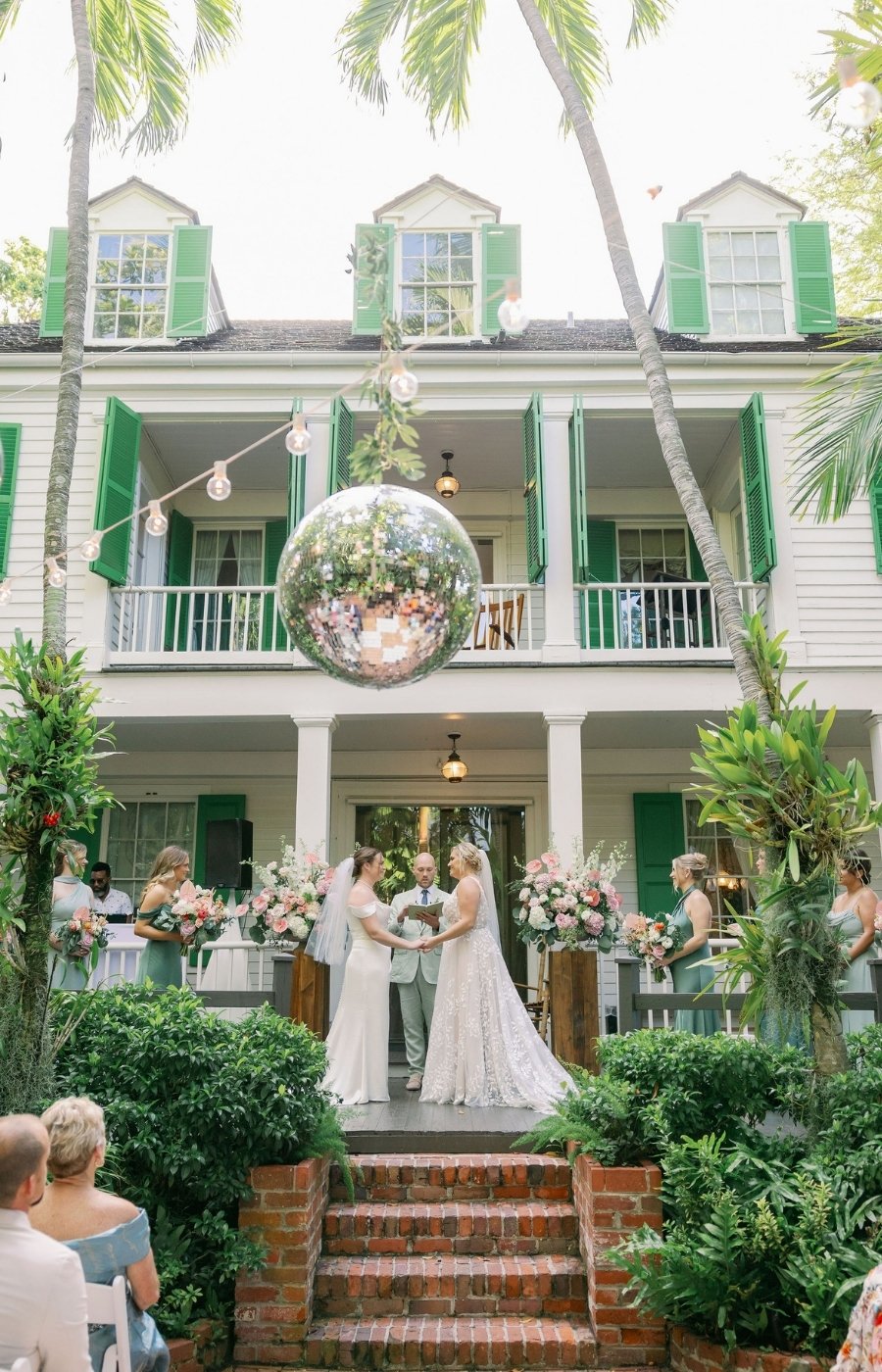 Two brides stand facing each other at an outdoor wedding ceremony in front of a large house with green shutters and a hanging mirrored sphere, surrounded by guests and greenery.