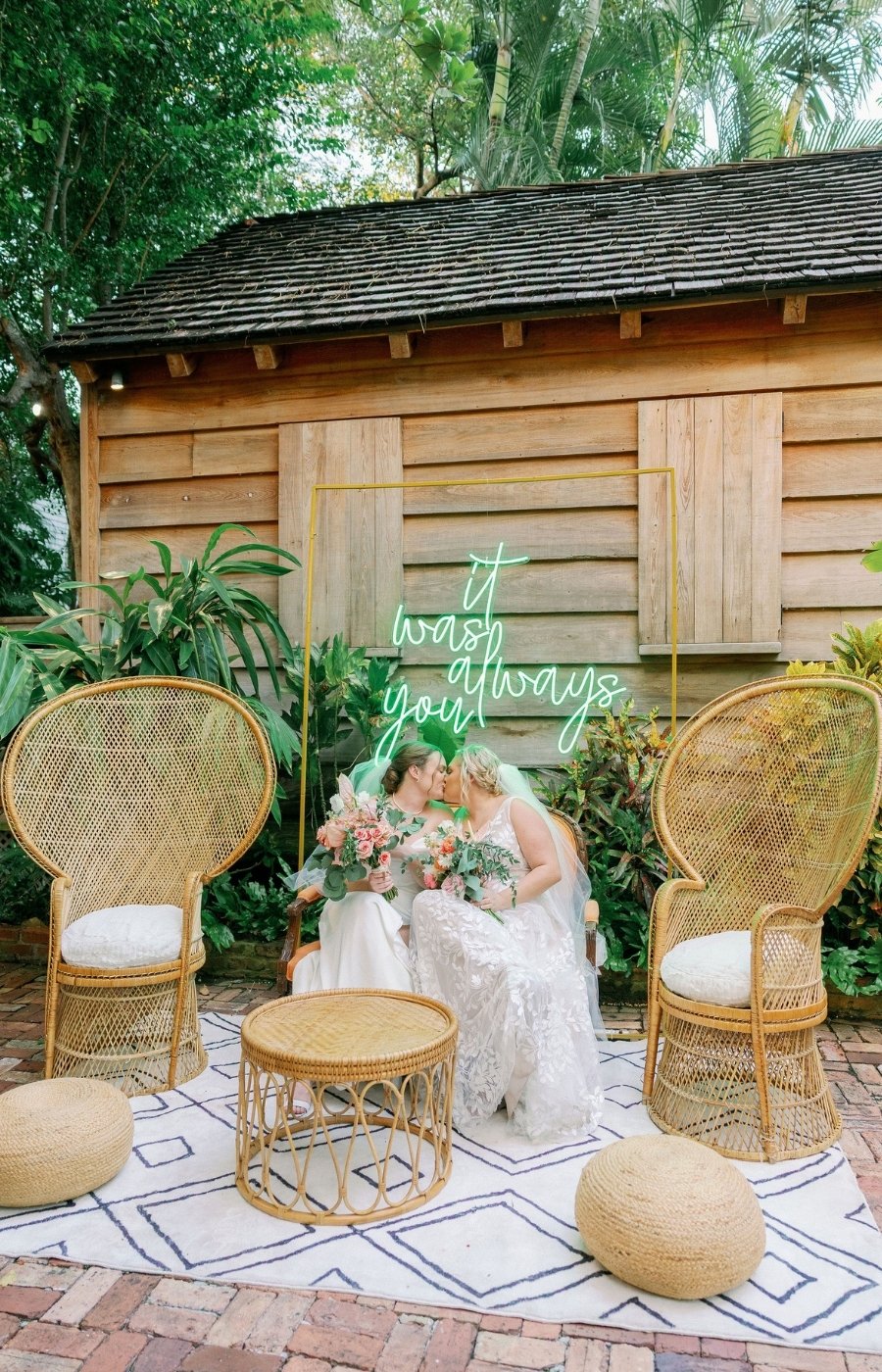 Two brides in wedding dresses sit together kissing and holding bouquets between wicker chairs, with a neon sign behind them reading "it was always you," in an outdoor garden setting.