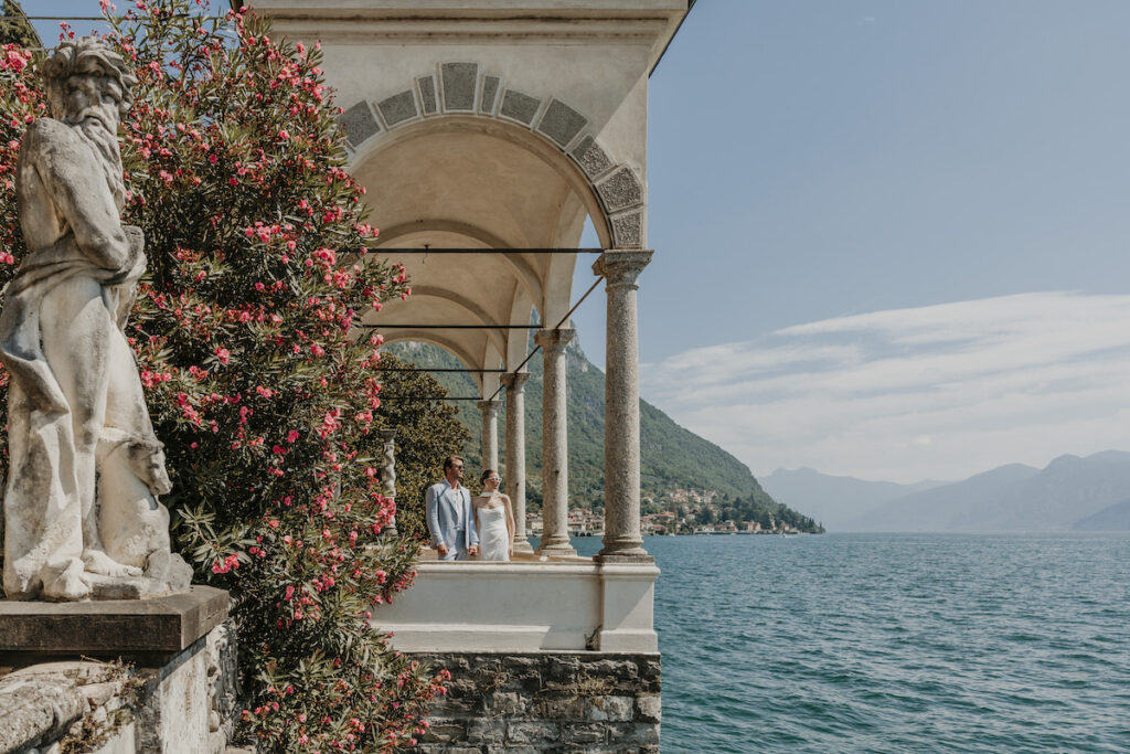 A couple stands under stone arches beside a flowering bush, overlooking a lake with mountains and a distant village in the background.