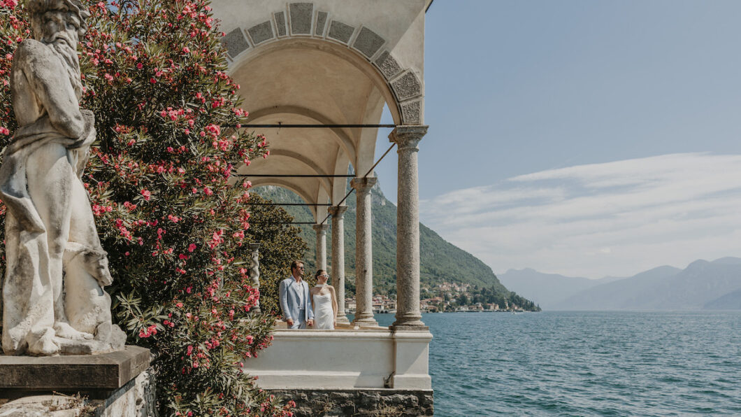 A couple stands under stone arches beside a flowering bush, overlooking a lake with mountains and a distant village in the background.