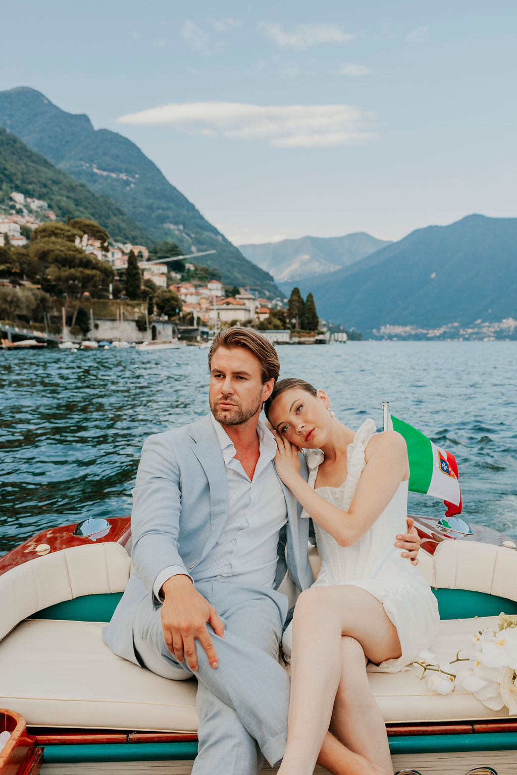 A couple sits on a boat with mountains and lakeside houses in the background; the woman rests her head on the man's shoulder.