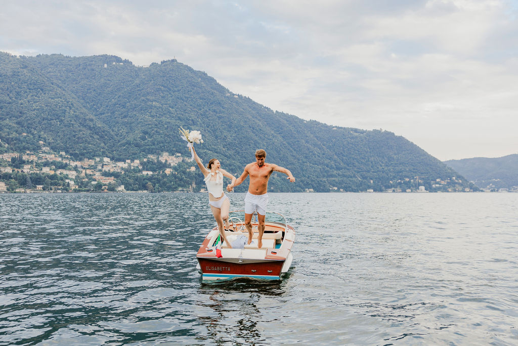 A woman in a white dress and a man in swim trunks stand on a small boat in a lake, with mountains and buildings in the background.