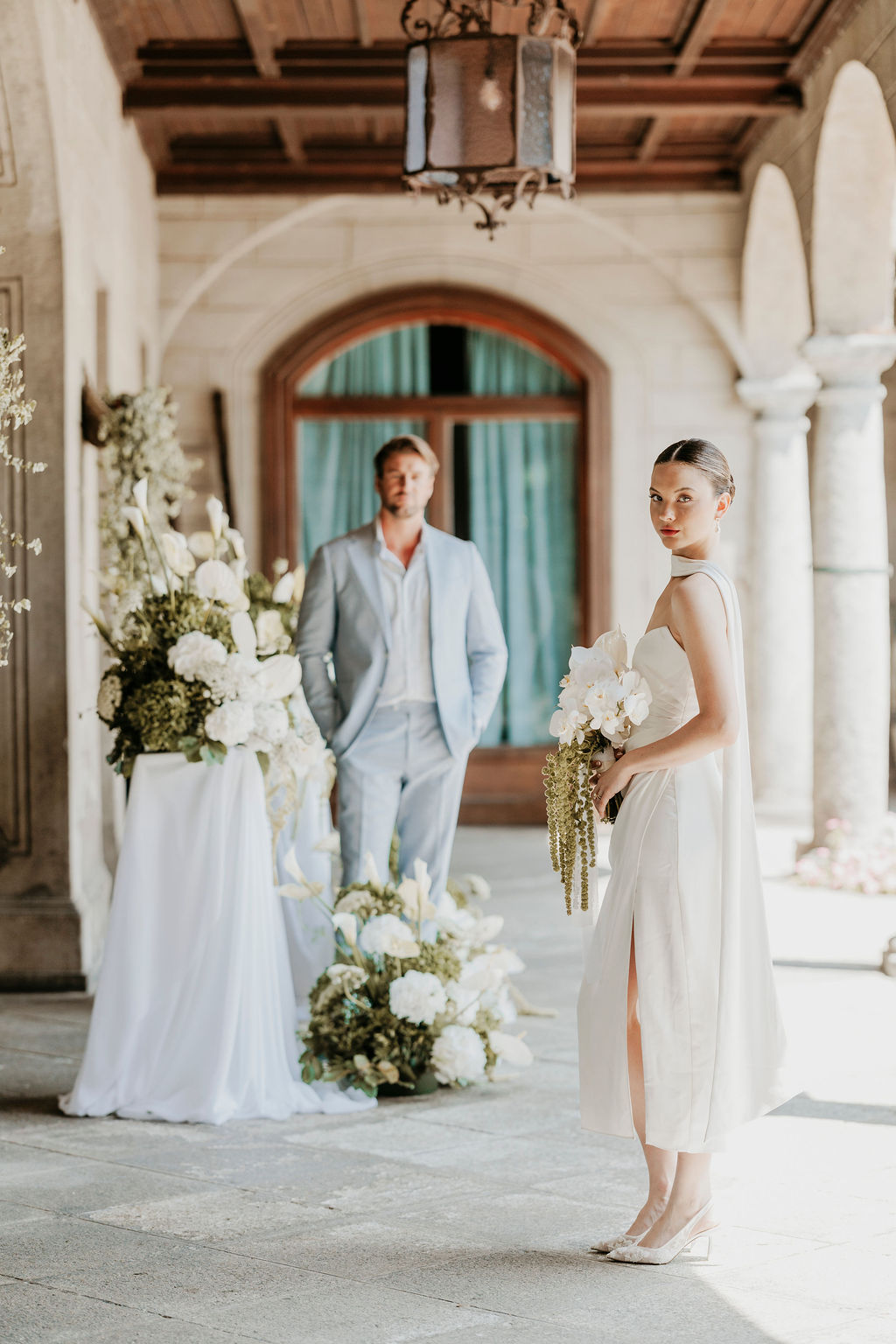 A woman in a white dress holds flowers and stands in the foreground, while a man in a light blue suit stands behind her in an elegant, arched hallway decorated with white flowers.