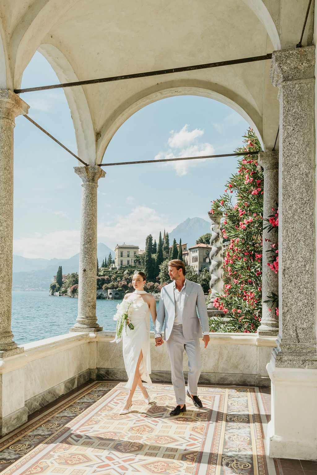 A couple dressed in formal attire walks hand in hand on a tiled terrace overlooking a lake and mountains, framed by stone arches and blooming flowers.