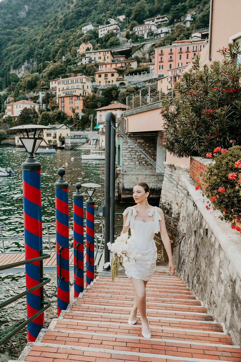 A woman in a white dress holding flowers walks up brick steps by a waterfront, with colorful buildings and greenery in the background.