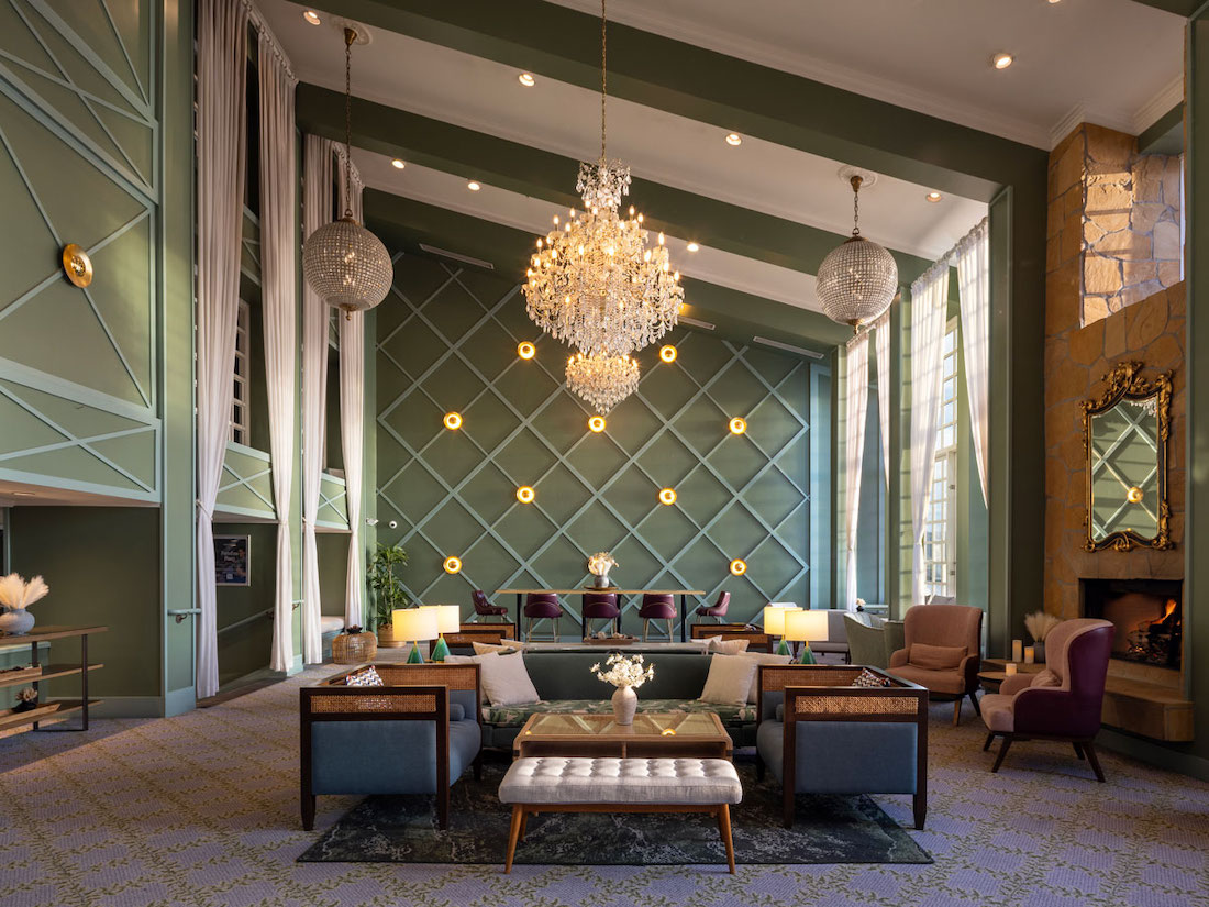 Elegant hotel lobby with high ceilings, large chandeliers, green paneled walls, multiple seating areas, and tall windows letting in natural light.