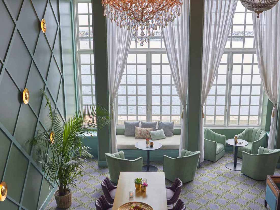 Elegant lounge with green armchairs, white tables, large windows with sheer curtains, a potted plant, and a chandelier hanging from the ceiling.
