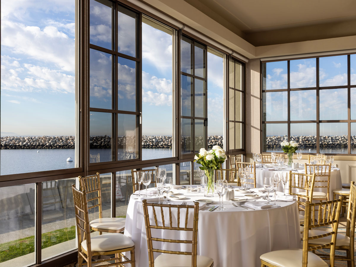 A round dining table with white linens and floral centerpiece is set in a sunlit room with large windows overlooking a waterfront and breakwater.