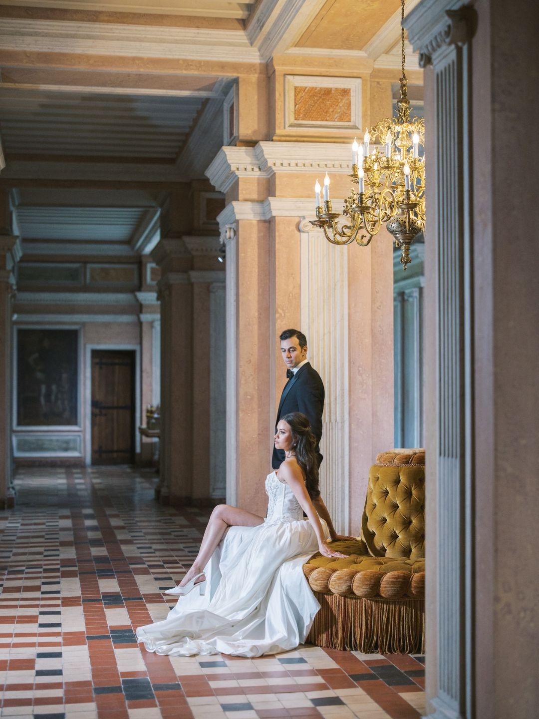 A woman in a white dress sits on a gold sofa, while a man in a suit stands behind her in an ornate hallway with checkered floors and a chandelier.