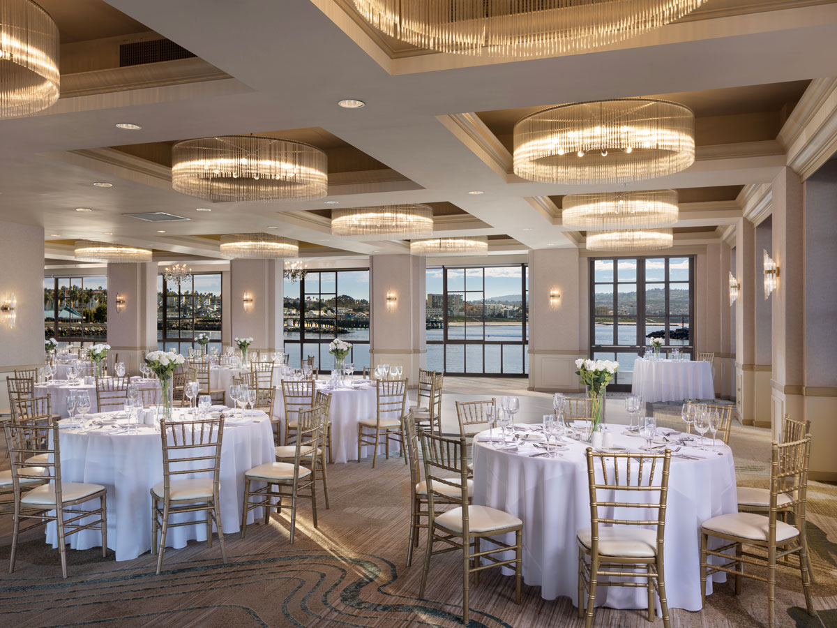 Elegant banquet hall with round tables set for an event, gold chairs, large windows overlooking water, and modern chandeliers overhead.